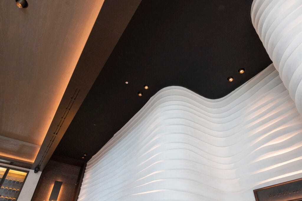 Zwart spanplafond met ingebouwde spots boven een organische witte designwand met indirecte LED-verlichting.